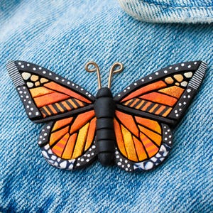 Bright Orange Monarch Butterfly pin on a denim jacket. Mosaic-style art brooch with bits of shine, texture and warm colors. Front angle