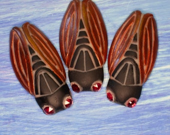 Puede incluir: Tres adornos decorativos con forma de insecto. Cada uno tiene alas marrones, cuerpo negro y ojos de gemas rojas. Los adornos están dispuestos sobre un fondo azul y blanco.