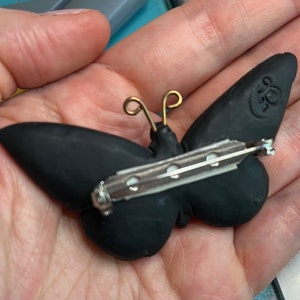 Shows the back of the brooch. Back is black with a secure bar clasp. Artist stamp is on the upper tip of right wing.