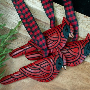 Photo shows 3 identical bright red cardinal ornaments hanging from red and black plaid ribbons.