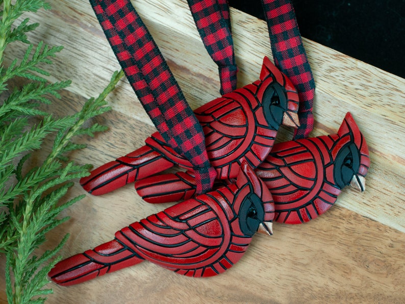 Photo shows 3 identical bright red cardinal ornaments hanging from red and black plaid ribbons.