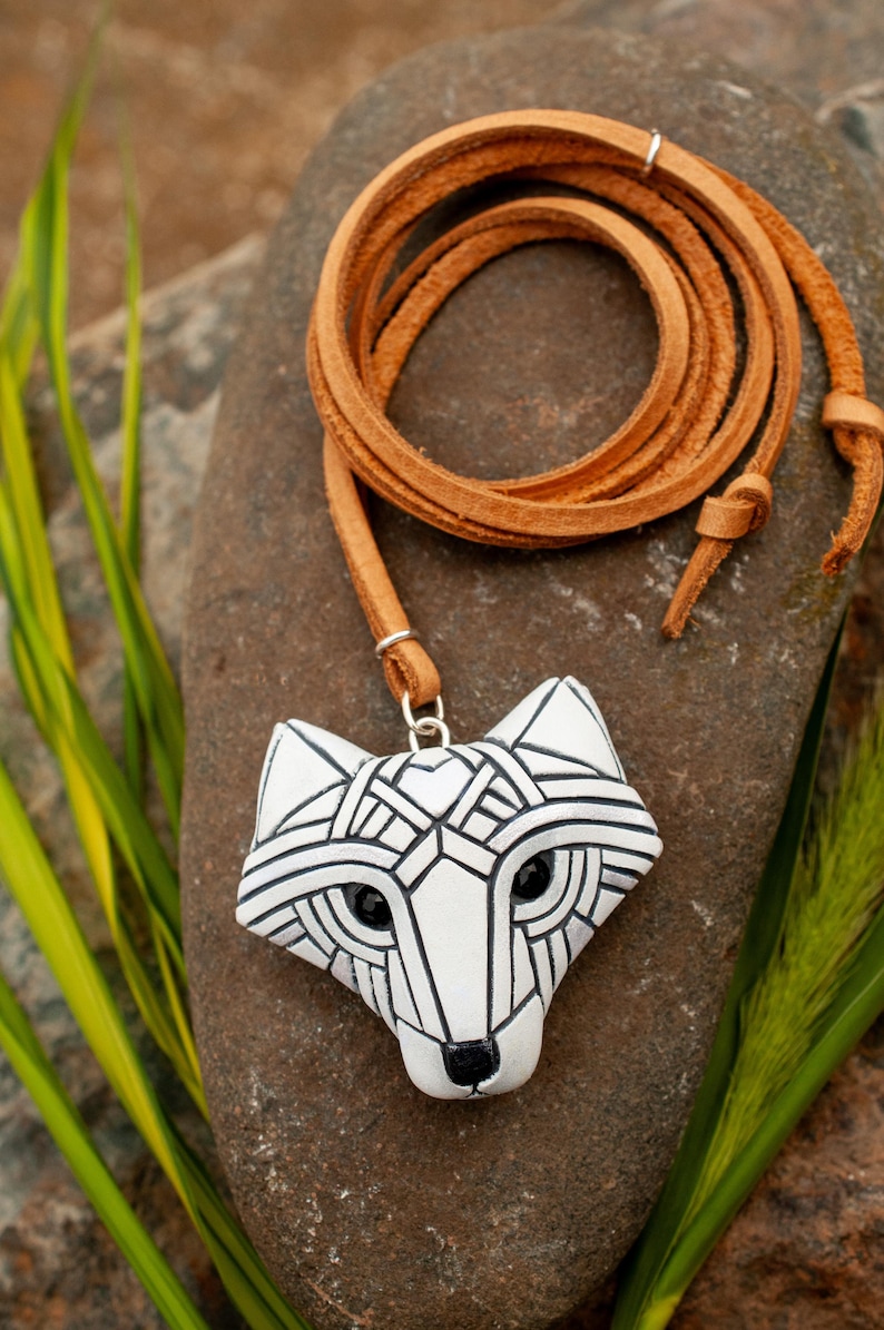 White Arctic fox necklace made in mosaic style is shown from the front. Pendant hangs from an adjustable tan leather cord. Eyes are sparkling black crystals. Fits in the palm of your hand. Made to look like an ancient amulet.