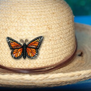 Bright Orange Monarch Butterfly pin on summer hat. Mosaic-style art brooch with bits of shine, texture and warm colors. Front angle