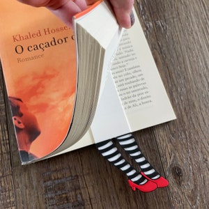 May include: A black and white striped bookmark with red shoes, shaped like a pair of legs, is sticking out of a book titled "O caçador d" by Khaled Hosseini.