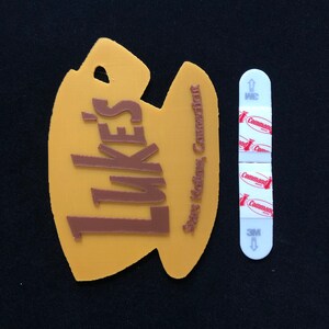 Puede incluir: Un letrero de pl&aacute;stico amarillo con forma de taza de caf&eacute; con el texto "Luke's" en letras marrones. El letrero tambi&eacute;n tiene el texto "Stars Hodiers, Connecticut" en letras marrones m&aacute;s peque&ntilde;as. El letrero est&aacute; sujeto a una tira adhesiva blanca de la marca Command.