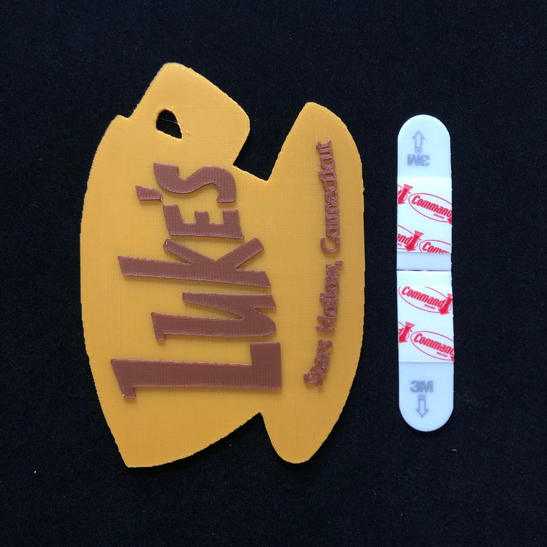 Puede incluir: Un letrero de pl&aacute;stico amarillo con forma de taza de caf&eacute; con el texto "Luke's" en letras marrones. El letrero tambi&eacute;n tiene el texto "Stars Hodiers, Connecticut" en letras marrones m&aacute;s peque&ntilde;as. El letrero est&aacute; sujeto a una tira adhesiva blanca de la marca Command.