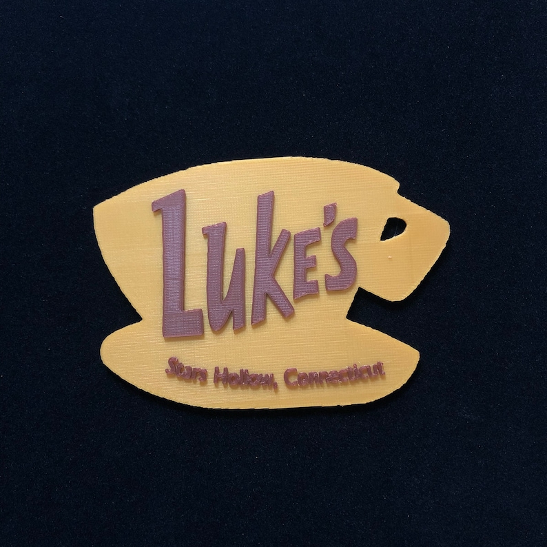 Puede incluir: Un letrero impreso en 3D de color amarillo y marr&oacute;n que dice "Luke's" con el texto "Stars Hollow, Connecticut" debajo. El letrero tiene la forma de una taza de caf&eacute; y un platillo.