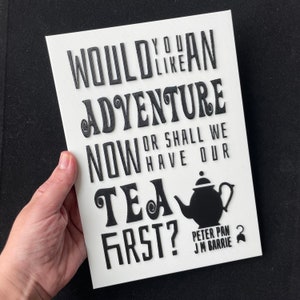 Peut inclure: Une toile blanche avec du texte noir : "Would you like an adventure now or shall we have our tea first ?" Un dessin de théière noire et le texte "Peter Pan J.M. Barrie" sont également présents.