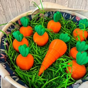 May include: A basket filled with orange carrot-shaped objects with green tops, nestled in green paper grass. The basket has a floral patterned lining and a white rope handle. One carrot is larger than the others.