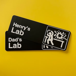 May include: Two black rectangular signs with white text. The top sign reads "Henry's Lab" and the bottom sign reads "Dad's Lab". The top sign has a white illustration of a person in a lab coat with their arms raised, standing in front of a table with a beaker exploding.