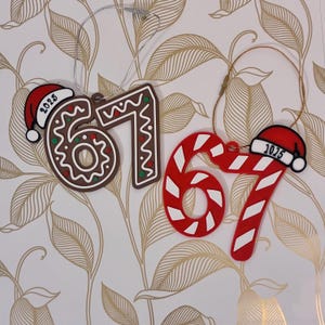 May include: Two Christmas tree decorations in the shape of the numbers 67. One is a gingerbread design, the other a red and white candy cane. Both have a Santa hat with the year 2025 and hanging strings.