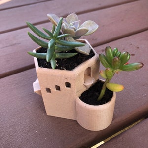 Puede incluir: Una maceta beige de estilo arquitectónico con tres suculentas. La maceta tiene un diseño tipo torre con aberturas arqueadas y una pequeña maceta redonda adjunta. Las suculentas son verdes y moradas.