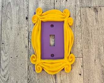 May include: A yellow plastic light switch cover shaped like the iconic frame from the TV show "Friends". The switch cover has a purple light switch plate.