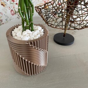 Op de afbeelding: Een decoratieve, geometrisch gevormde plantenbak in bronskleur, gevuld met witte stenen en groene bamboestengels. Een draadsculptuur staat op de achtergrond. De plantenbak staat op een grijze ondergrond.