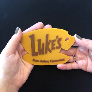 Puede incluir: Un letrero amarillo con las palabras "Luke's" en letras marrones. El letrero tambi&eacute;n dice "Stars Hollow, Connecticut" en letras marrones m&aacute;s peque&ntilde;as.