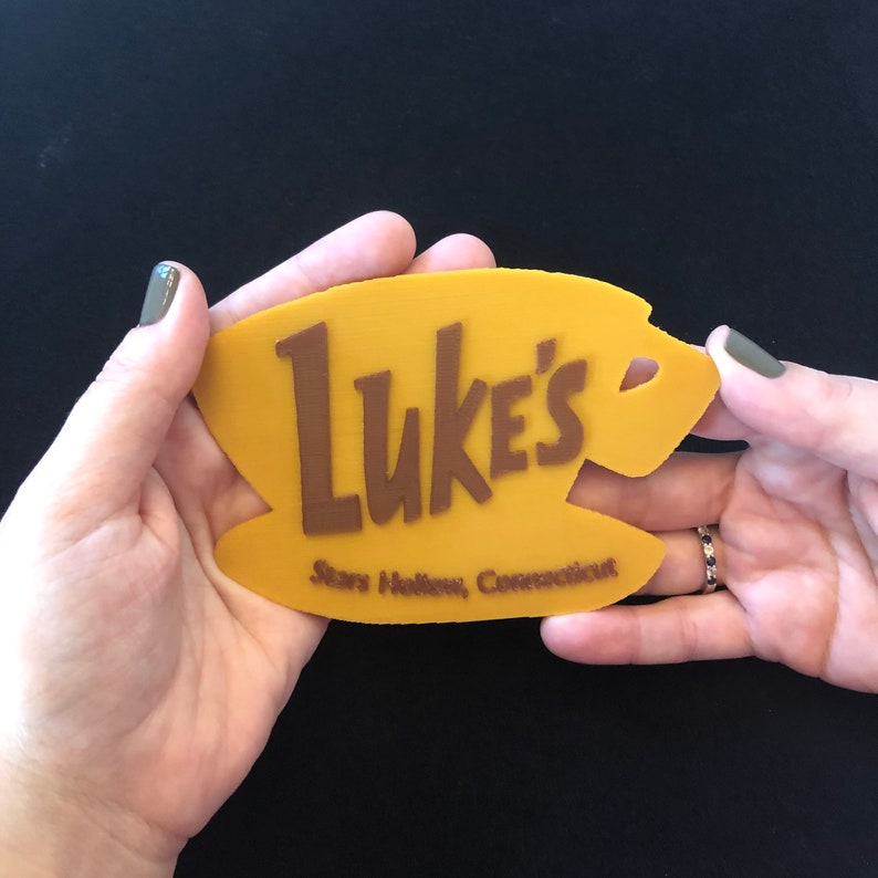 Puede incluir: Un letrero amarillo con las palabras "Luke's" en letras marrones. El letrero tambi&eacute;n dice "Stars Hollow, Connecticut" en letras marrones m&aacute;s peque&ntilde;as.
