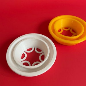Pode incluir: Dois objetos impressos em 3D sobre um fundo vermelho. Um é branco, o outro amarelo. Ambos têm um design circular com uma forma de tigela central e pequenos recortes curvos. O objeto branco é maior que o amarelo.