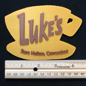Puede incluir: Un letrero amarillo impreso en 3D con las palabras "Luke's" en texto marr&oacute;n y las palabras "Stars Hollow, Connecticut" en texto marr&oacute;n m&aacute;s peque&ntilde;o. El letrero tiene forma de taza de caf&eacute;.