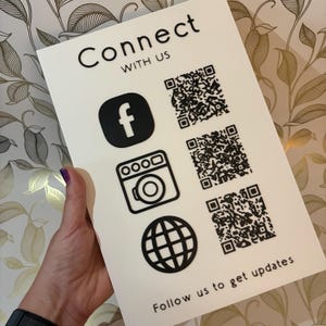 May include: A white sign with the words "Connect with Us" in black. The sign features a Facebook logo, a camera icon, and a globe icon, along with three QR codes. The text "Follow us to get updates" is at the bottom.