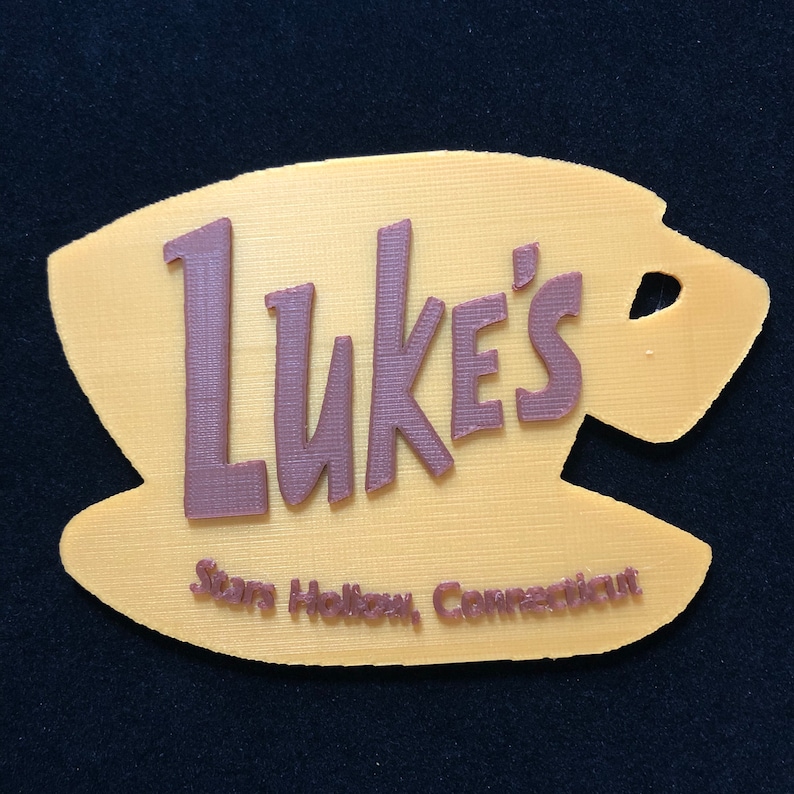 Puede incluir: Un letrero amarillo impreso en 3D con forma de taza de caf&eacute; con el texto "Luke's" en letras marrones. El texto "Stars Hollow, Connecticut" tambi&eacute;n est&aacute; impreso en letras marrones debajo del texto principal.