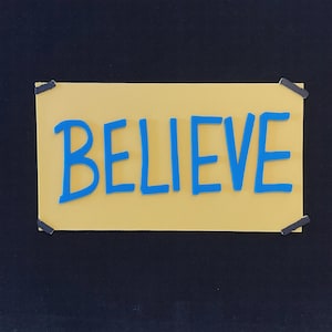 May include: A yellow rectangular sign with the word "BELIEVE" in blue letters. The sign is attached to a black surface with four small black pieces of tape at each corner.