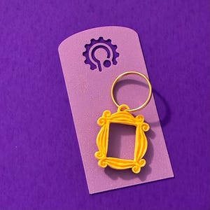Könnte beinhalten: Ein gelber schlüsselanhänger in Rahmenform mit einem goldfarbenen Ring, befestigt an einem lila Anhänger. Der Rahmen hat eine quadratische Öffnung und dekorative Wirbel. Der Anhänger hat oben einen zahnradförmigen Ausschnitt.