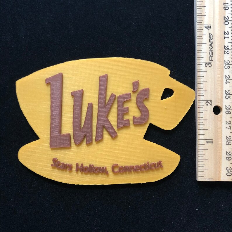 Puede incluir: Un letrero impreso en 3D de color amarillo y marr&oacute;n que dice "Luke's Stars Hollow, Connecticut". El letrero tiene la forma de una taza de caf&eacute;.