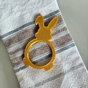 Könnte beinhalten: Ein goldfarbener, hasenförmiger Serviettenring. Der Ring besteht aus einem strukturierten Material und liegt auf einem gestreiften beigefarbenen und braunen Tuch. Der Hase hat lange Ohren und eine kreisförmige Öffnung für eine Serviette.