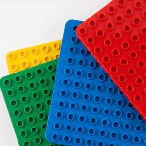 Op de afbeelding: Een close-up van kleurrijke speelgoedbouwstenen in rood, blauw, groen en geel. Elke blok heeft een rechthoekige vorm met cirkelvormige noppen op het bovenoppervlak. De blokken zijn in verschillende hoeken gerangschikt, wat hun levendige kleuren en ontwerp laat zien.