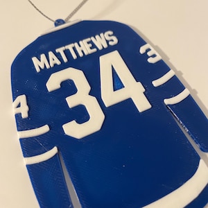 May include: Blue and white jersey-shaped ornament with the name "MATTHEWS" and the number "34" in white. The ornament is designed to resemble a hockey jersey, with white stripes on the sleeves.