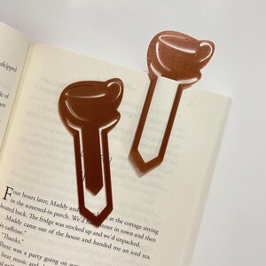 May include: Two brown coffee cup-shaped bookmarks are placed on the pages of an open book. The bookmarks are made of a solid material and have a cutout design. The book is open to a page of printed text.