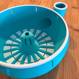 Pode incluir: Um espremedor de citrinos turquesa e branco. O espremedor tem um alargador central com nervuras radiais e uma bacia circundante. Um pequeno bico estende-se da lateral. O design é moderno e funcional, adequado para uso na cozinha.