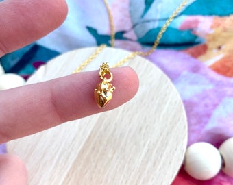 May include: A gold necklace with a small, detailed anatomical heart pendant. The pendant hangs from a fine gold chain. The necklace is shown on a wooden surface, with a colourful floral background.