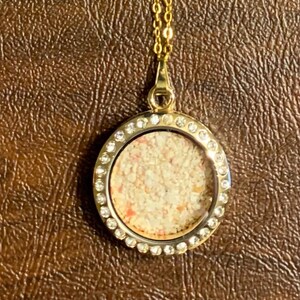 May include: Gold necklace with a round pendant featuring a clear glass insert filled with sand and surrounded by small clear rhinestones.