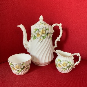May include: A white porcelain tea set with a floral design. The set includes a teapot, a creamer, and a teacup. The teapot has a spiral design and gold trim. The floral design features white and yellow flowers with green leaves.