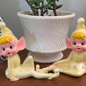May include: Two vintage ceramic elf figurines in a relaxed pose, with yellow outfits, pointy hats, and large pink ears. The elves have rosy cheeks, red lips, and blonde hair. A white, swirled planter with a green plant sits behind them.