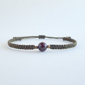 May include: A dark green braided cord bracelet with a single dark red gemstone bead.