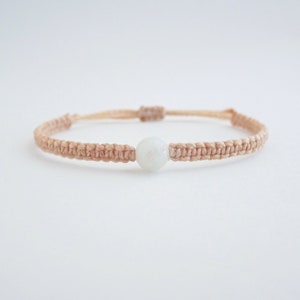 May include: A delicate bracelet made with a light brown cord and a single white gemstone bead.