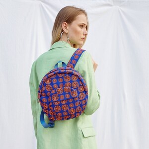 May include: A woman wearing a light green blazer and white pants is wearing a round blue and orange patterned backpack. The backpack has a circular design with orange swirls on a blue background.