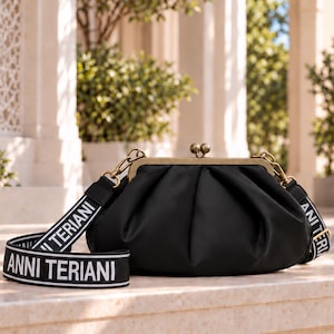 May include: A black clutch bag with a gathered, rounded silhouette and a brass-toned metal kiss-lock frame. It features a detachable black shoulder strap with the white text ANNI TERIANI printed repeatedly along the length of the fabric strap.