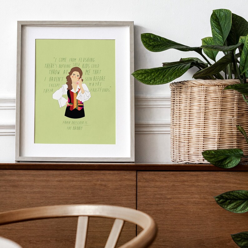 May include: Framed art print featuring a cartoon illustration of Fran Drescher from The Nanny, with a quote from the show. The artwork has a light green background and is in a gray frame. The print is displayed on a wooden cabinet.