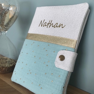 Peut inclure: Un livre pour bébé bleu clair avec des points de paillettes dorés et une couverture blanche avec un ruban doré et le nom "Nathan" brodé dessus.