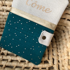 Op de afbeelding: Een tealblauwe stoffen boekomslag met gouden stippen en een gouden lint. De omslag heeft een witte flap met een knoopsluiting en het woord "Come" is in gouden draad geborduurd op de voorkant.