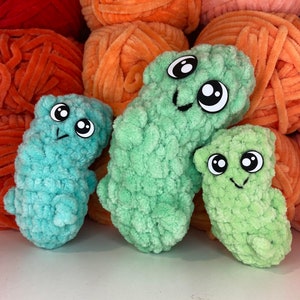 May include: Three crocheted plush toys shaped like a worm with googly eyes and smiling faces. The toys are green and turquoise.