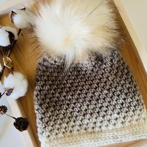 May include: A brown and white ombre crochet beanie with a fluffy white pom pom.