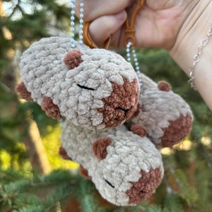 May include: Three crocheted capybara keychains. Each keychain features a light brown body with darker brown accents and closed black eyes. The keychains are attached to a brown clasp with a small chain.