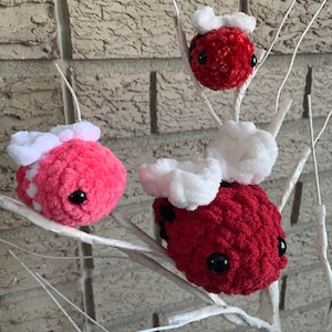 May include: Three crocheted ladybug ornaments with white wings and black eyes. The ladybugs are hanging on a white branch.