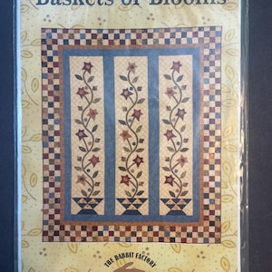 May include: A quilt pattern titled "Baskets of Blooms" featuring a floral design. The quilt design has three vertical panels with blue backgrounds and floral vines. The border has a checkered pattern and a yellow background with leaf designs. The Rabbit Factory logo is at the bottom.