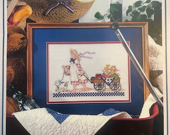 May include: A magazine cover for Cross Stitch, featuring a girl with a wagon full of produce. The magazine cover is red, white, and blue with the text "Cross Stitch" in large, bold letters. The magazine is celebrating 10 years of excellence.