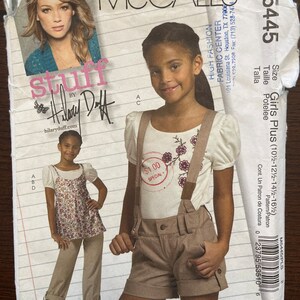 May include: A sewing pattern for a girl's plus size outfit. The pattern includes a white t-shirt with a floral design and a pair of brown shorts with suspenders. The pattern is from McCall's and is size 102-122-142-162.